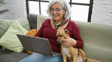 Gülümseyen, gri saçlı kadın videosu. Oturma odasında kucağında bir köpekle arıyor.