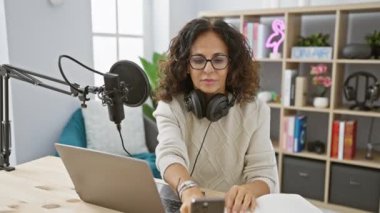 Orta yaşlı İspanyol bir kadın iyi donanımlı bir radyo stüdyosunda podcast sunuyor, notlar alıyor ve izleyicileriyle iletişim kuruyor..