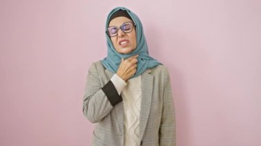Orta yaşlı, tesettürlü bir kadın, ağrıyan boynunu tutuyor, soğuk algınlığı ve enfeksiyonu pembe izole bir arka planda hissediyor.
