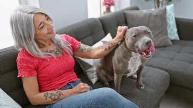 Evdeki kanepede oturan neşeli orta yaşlı kadın, sevgili gri saçlı köpeğiyle oynuyor, gülüyor ve saf mutluluğu ifade ediyor.