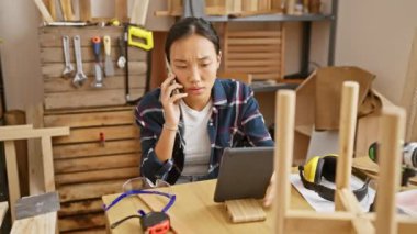 Genç Asyalı bir kadın, iyi donanımlı bir marangoz atölyesinde tablet kullanırken telefonla konuşuyor..