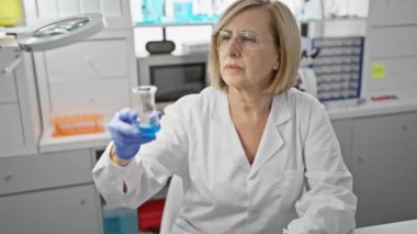 Laboratuvarda olgun bir kadın bilim adamı mavi sıvıyla bir şişeyi inceliyor, profesyonelliği ve konsantrasyonu resmediyor.