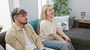 Genç bir adam ve kadın birlikte gri bir kanepede, parlak bir oturma odasında oturuyor, şefkat ve gülümseme sergiliyorlar..