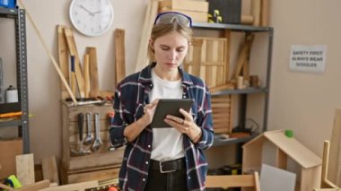 Tabletle odaklanmış bir kadın marangozluk atölyesinde ahşap işçiliği aletleri ve kerestelerle dolu duruyor..