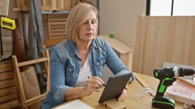 Olgun bir kadın ahşap atölyesinde tablet kullanıyor. Etrafı aletler ve güvenlik ekipmanlarıyla çevrili..