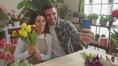 Çiçekçide selfie çeken erkek ve kadın çiçekçiler. Etrafı bitkilerle ve renkli çiçeklerle çevrili..