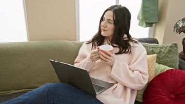Rahat bir kadın evdeki rahat bir kanepede kahve ve laptoptan hoşlanır.