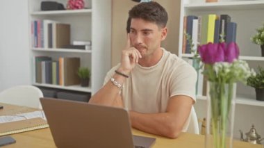 Yakışıklı İspanyol adam, modern bir oturma odasında dizüstü bilgisayar ve çiçeklerle düşünüyor.