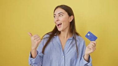 Kredi kartı olan neşeli, genç bir İspanyol kadın sarı arka planda izole edilmiş bir şekilde baş parmağını kaldırıyor.