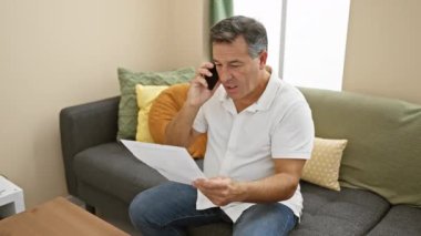 Oturma odasında elinde belge tutarken telefonla konuşan olgun bir adam endişe ve konsantrasyonunu dile getiriyor..