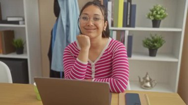 Gözlüklü neşeli genç bir kadın, ev ofisinde oturuyor, kameraya gülümsüyor, etrafı bitkiler ve kitaplarla çevrili..