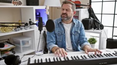 Yakışıklı, orta yaşlı, gri saçlı, piyano çalan ve modern bir müzik stüdyosunda şarkı söyleyen bir adam..