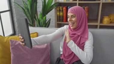 Tesettürlü neşeli genç bir kadın evdeki video çağrısının tadını çıkarıyor. Oturma odasında mutluluk ve rahatlığı ifade ediyor.