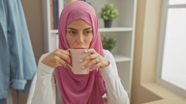 Kafiyeli bir kadın evin içinde bitki ve pencere pervazıyla çay yudumluyor..