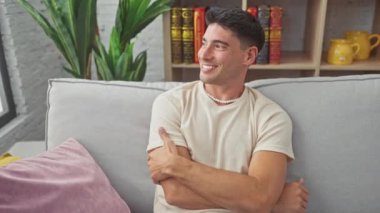 Neşeli, genç, İspanyol bir adam rahatça başını çeviriyor, oturma odasındaki kanepede oturuyor. Yakışıklı bir yetişkinin doğal gülümsemesini, güvenen yüz ifadesini, evdeki profilini gösteren bir fotoğraf..