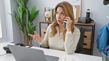 Modern bir ofis ortamında dizüstü bilgisayarla çalışan profesyonel bir İspanyol kadın telefonda konuşuyor..