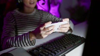 Klavye ve neon ışıklarıyla karanlık bir oyun odasında akıllı telefondan mesaj atan gülümseyen İspanyol kadın.