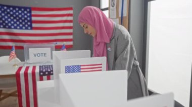 Elinde tesettür ve seçmen etiketi olan gülümseyen bir kadın bizimle birlikte bir oy kullanma merkezinde Amerikan demokratik sürecine katılıyor..