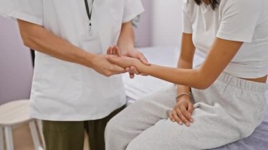 Laboratuvar önlüklü bir adam, parlak bir rehabilitasyon kliniğinde oturan bir kadının elini inceliyor. Sağlık hizmetleri ve profesyonel terapi hizmetleri sunuyor..