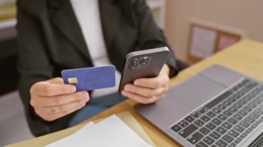 Kredi kartı ve akıllı telefonu elinde tutan İspanyol kadın bir ofis masasında dizüstü bilgisayarı ve kağıtları olan..