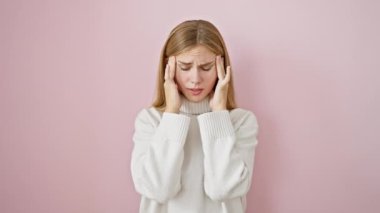 Stresli ve çarpıcı, güzel sarışın kız migren ağrısı çekiyor. Pembe arka planda, acının ağırlığı altında tek başına duruyor.