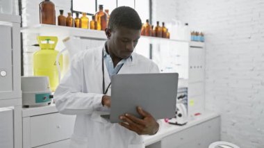 Modern kimya laboratuvarındaki bir dizüstü bilgisayardaki verileri analiz eden Afrikalı Amerikalı erkek bilim adamı..