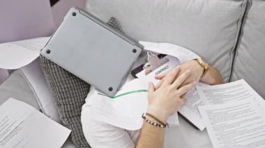 Evrak işleri ve bilgisayarla çevrili yetişkin bir adam, kapalı bir odada gri bir kanepede otururken...