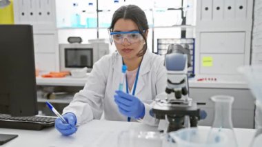 Odaklanmış bir İspanyol kadın bilim adamı laboratuarda bir örnek inceliyor, mikroskobun yanında sonuçlar yazıyor..