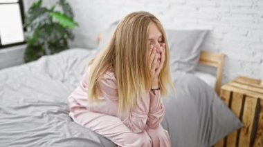 Genç sarışın bir kadın yatak odasında bir yatakta oturuyor, elleriyle yüzünü kapatıyor, üzgün ya da üzgün görünüyor..