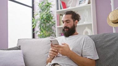 Modern oturma odasındaki kanepede dinlenirken akıllı telefon kullanan yakışıklı sakallı adam.