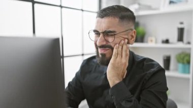 İşyerinde otururken yanağına dokunan sakallı bir adam modern ofis ortamında diş ağrısına işaret ediyor..