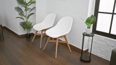 Sırt çantalı sakallı adam modern bir bekleme odasında oturuyor, bitkilerin ve minimalist sandalyelerin yanında bir defter tutuyor..