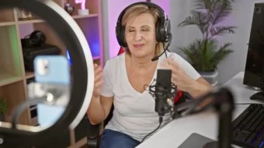 Başarılı orta yaşlı sarışın flamalı kadın, eğlenceli bir zaferi kutluyor. Kasetteki neşeli bir gülümsemeyle karanlık oyun odasında..