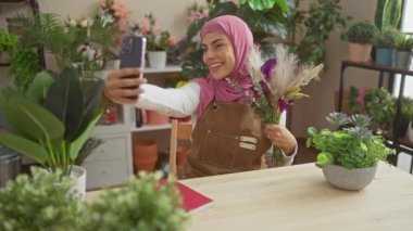 Başörtüsü içinde gülümseyen kadın, kapalı bir odada akıllı bir telefonla selfie çekiyor.
