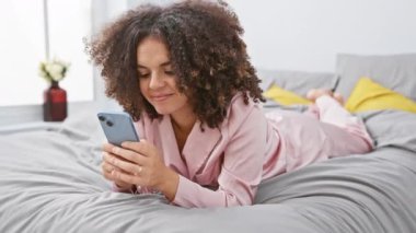 Yatakta kıvırcık saçlı neşeli İspanyol kadın zaferini kutlarken mutlu bir şekilde gülümsüyor ve yatak odasında akıllı telefon kullanıyor.