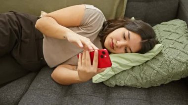 İspanyol kadın evdeki kanepede dinleniyor, rahat ve rahat bir şekilde akıllı telefon kullanıyor.