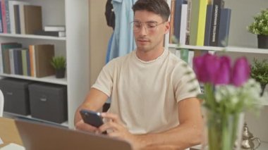 Genç bir İspanyol adam, modern bir ofis ortamında akıllı telefon kullanarak uzaktan çalışma ve teknoloji kullanımını resmederken gülümsüyor..