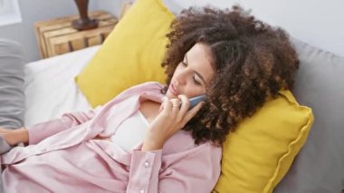 Kıvırcık saçlı, genç bir İspanyol kadın evinde rahat rahat yastıklara yaslanarak akıllı bir telefonla konuşuyor..