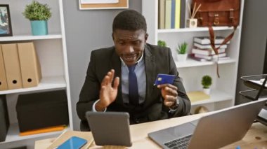 Takım elbiseli Afrikalı bir adam, modern bir ofiste video görüşmesi yaparken kredi kartını inceliyor..