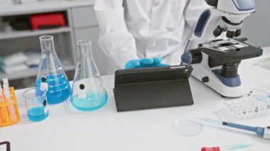 Laboratuvar çalışma alanında mikroskop ve tabletle mavi sıvıyı analiz eden bilim kadını.