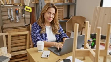 Neşeli genç Latin kadın kendine güveni tam, marangozluk masasında otururken serin bir gülücük sallıyor, atölyesinde bir neşe ve başarı portresi.