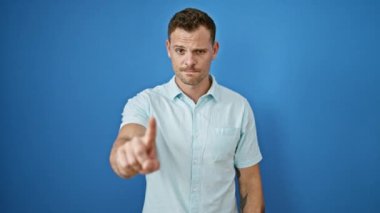 Sakallı hoşnutsuz bir genç adam, dışarıda mavi duvar arka planına karşı baş parmak kaldırıyor..
