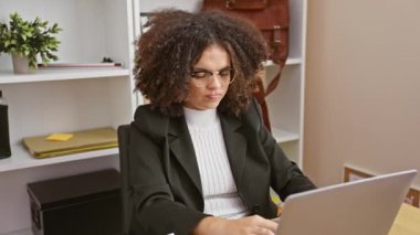 Ofisinde dizüstü bilgisayarla çalışırken boynu ağrıyan kıvırcık saçlı İspanyol bir kadın.