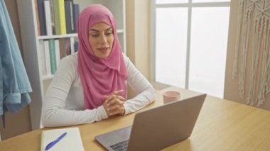Kendine güvenen, tesettürlü, tesettürlü genç bir kadın. Ciddi bir ifadeyle parmağını sana doğrultuyor. Dizüstü bilgisayarında, oturma odasının arka planında.