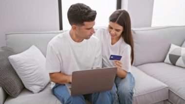 Kredi kartı ve dizüstü bilgisayarı olan gülümseyen bir çift modern bir oturma odasındaki kanepede rahatça alışveriş yapıyor..