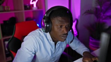 Kulaklık takan odaklanmış siyah bir adam geceleri neon ışıklı bir oyun odasında bilgisayarla meşgul oluyor..