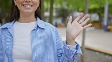 Çizgili tişörtlü gülümseyen genç İspanyol kadın elini güneşli bir parkta sunuyor.