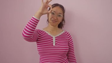 Neşeli, genç İspanyol bir kadın çizgili bir tişörtle duruyor, mutlu bir şekilde eliyle işaret yapıyor, gözünün üzerinden pembe bir arka planda parmaklarının arasından bakıyor..