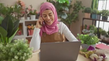 Tesettürlü gülümseyen bir kadın, bitkilerle dolu bir ofiste dizüstü bilgisayar kullanırken telefonla konuşur..
