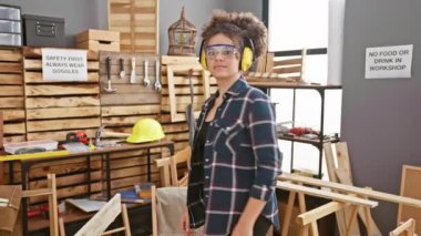 Kıvırcık saçlı, gülümseyen genç bir kadın bir marangoz atölyesinde güvenlik gözlüğü ve kulak koruyucu takıyor.